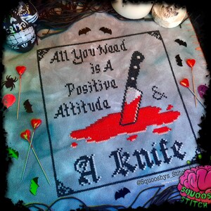 May include: Cross stitch pattern with a black and white design that reads "All You Need Is A Positive Attitude & A Knife". The design is on a blue background with red blood splatters and a knife.