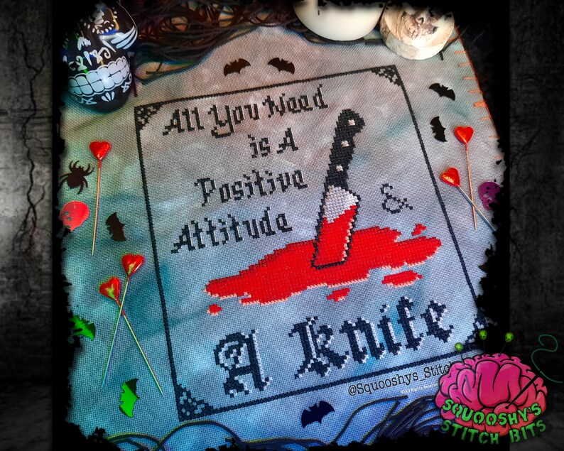 May include: Cross stitch pattern with a black and white design that reads "All You Need Is A Positive Attitude & A Knife". The design is on a blue background with red blood splatters and a knife.