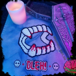 May include: Cross-stitch embroidery on blue fabric featuring a pair of fangs with dripping blood, the text "GLEH!" and two skull icons. The embroidery is framed by a wooden coffin with a rose design.