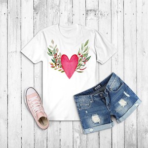 May include: White t-shirt with a red heart surrounded by green leaves and red berries. The heart is painted in a watercolor style. The t-shirt is laid flat on a white wooden surface with a pair of blue denim shorts and pink sneakers.