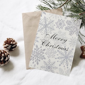 K&ouml;nnte beinhalten: Eine wei&szlig;e Karte mit einem Schneeflockenmuster und dem Text "Merry Christmas" in schwarzer Schrift. Die Karte liegt auf einer wei&szlig;en Oberfl&auml;che mit einem braunen Umschlag, zwei Tannenzapfen und einem Lebkuchen in Sternform mit einem Schneeflockenmuster.