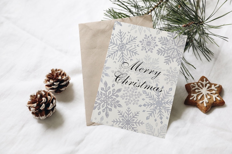 K&ouml;nnte beinhalten: Eine wei&szlig;e Karte mit einem Schneeflockenmuster und dem Text "Merry Christmas" in schwarzer Schrift. Die Karte liegt auf einer wei&szlig;en Oberfl&auml;che mit einem braunen Umschlag, zwei Tannenzapfen und einem Lebkuchen in Sternform mit einem Schneeflockenmuster.
