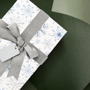 May include: A white gift box with a silver ribbon and a snowflake pattern. The box has a blank white tag attached to the ribbon.