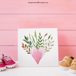 May include: A white card with a watercolor painting of a pink heart surrounded by green and brown leaves. Two pairs of baby shoes, one pink and one brown, are on either side of the card on a white surface.