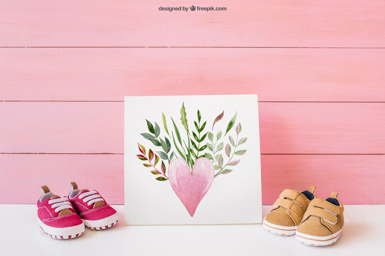 May include: A white card with a watercolor painting of a pink heart surrounded by green and brown leaves. Two pairs of baby shoes, one pink and one brown, are on either side of the card on a white surface.
