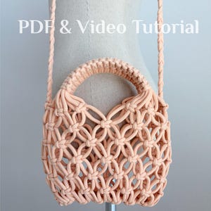 Puede incluir: Un bolso de macramé color melocotón con un intrincado patrón de nudos en celosía, un asa superior redondeada y una correa trenzada. El texto "PDF & Video Tutorial" aparece en la parte superior, indicando contenido instructivo.