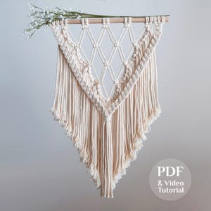 Op de afbeelding: Crèmekleurige macramé wandhanger met een geometrisch ruitpatroon en franjes. De macramé is opgehangen aan een houten deuvel en versierd met witte bloemen. De afbeelding bevat de tekst "PDF & Video Tutorial".
