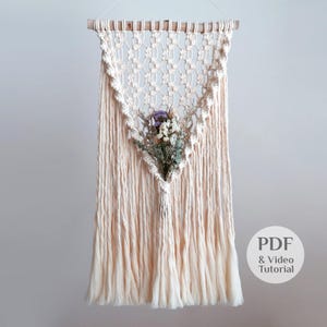 May include: Cream-colored macrame wall hanging with a triangular design, featuring a bouquet of dried flowers. The piece is suspended from a wooden dowel and includes a fringe at the bottom. The text "PDF & Video Tutorial" is visible.