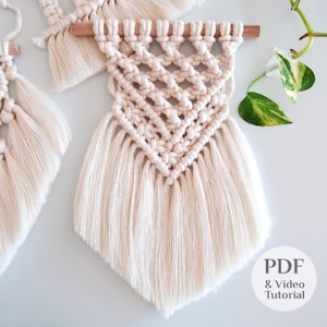 May include: Cream-colored macrame wall hanging with a geometric diamond pattern and fringe. The macrame is attached to a natural wooden dowel. The image also includes a PDF & Video Tutorial graphic.
