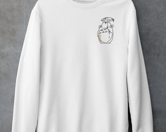 May include: White crewneck sweatshirt with a small embroidered design of a cartoon character under an umbrella. The design is outlined in black with tan and gray accents. The sweatshirt is hanging on a copper-colored hanger against a gray wall.