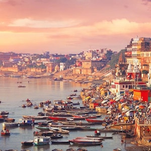 Może przedstawiać: Panoramiczny widok na panoramę miasta wzdłuż rzeki z wieloma łodziami. Miasto jest zbudowane na zboczu i ma wiele kolorowych budynków i świątyń. Rzeka jest spokojna, a niebo jest w delikatnym różowym i pomarańczowym kolorze.