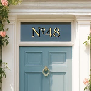 May include: A teal front door with gold hardware and the number "N&deg;48" above. The door is surrounded by white trim and climbing roses with pink blooms. A classic and inviting entrance.