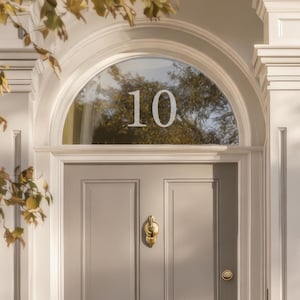 Pode incluir: Uma porta da frente cinza claro com o número 10 numa janela semicircular. A porta tem uma aldrava e uma ranhura para correspondência em latão. A porta é emoldurada por molduras brancas e uma vista parcial de uma árvore com folhas amarelas.