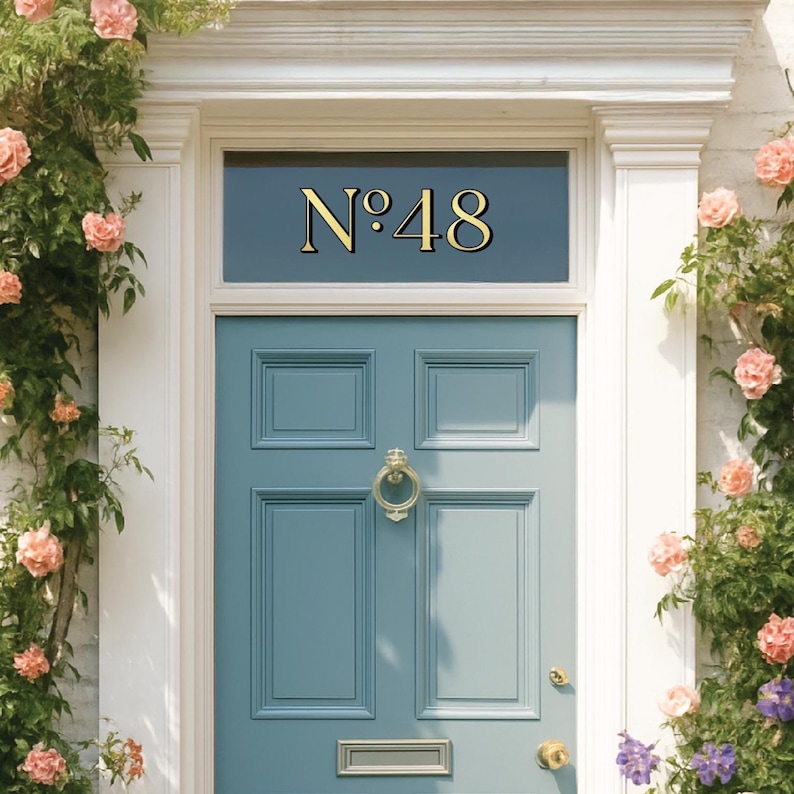 May include: A teal front door with gold hardware and the number "N&deg;48" above. The door is surrounded by white trim and climbing roses with pink blooms. A classic and inviting entrance.