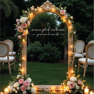 May include: A decorative wedding arch made of a gold-painted wooden frame with a mirror inside. The arch is decorated with pink and white flowers and string lights. The text "We're so glad you're here - your seat awaits -" is written on the mirror.