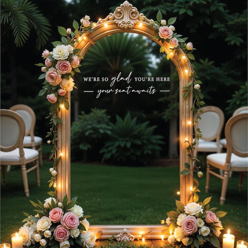 May include: A decorative wedding arch made of a gold-painted wooden frame with a mirror inside. The arch is decorated with pink and white flowers and string lights. The text "We're so glad you're here - your seat awaits -" is written on the mirror.