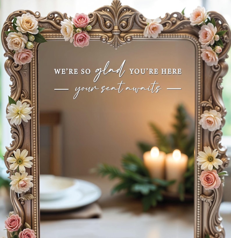 May include: A gold ornate mirror with a white inscription that reads "We're so glad you're here - your seat awaits -." The mirror is decorated with artificial flowers in shades of pink and white.
