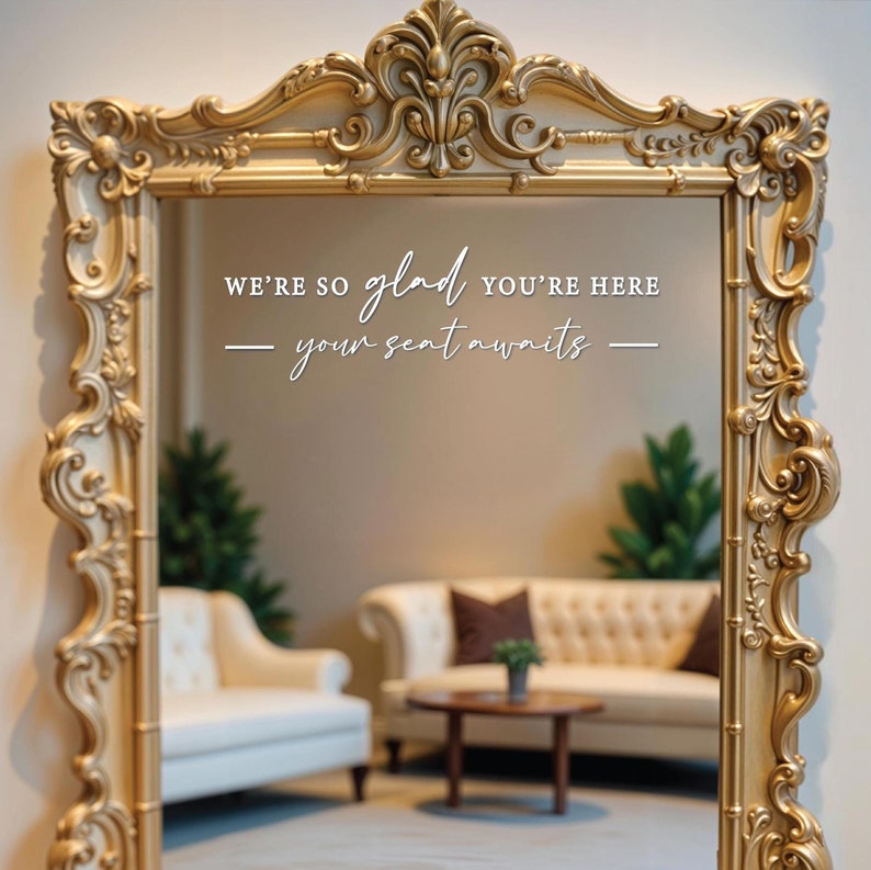 May include: A gold ornate mirror with the text "We're so glad you're here - your seat awaits -" reflected in the glass. The mirror is reflecting a room with two white sofas and a wooden coffee table.
