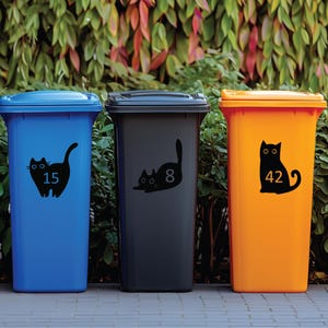 Könnte beinhalten: Drei blaue, schwarze und orangefarbene Recyclingbehälter mit schwarzen Katzenaufklebern und den Nummern 15, 8 und 42.