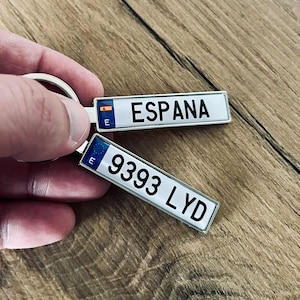 May include: Two rectangular keyrings with a silver border. One reads "ESPANA" with the Spanish flag and the letter "E". The other reads "9393 LYD" with the letter "E" and a blue background.