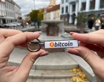 May include: A silver keychain with a white rectangular tag that says "bitcoin" in black text. The bitcoin symbol is orange and is located on the left side of the tag.