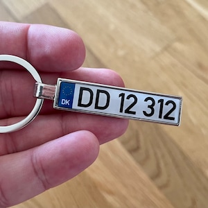May include: Silver keyring with a blue and yellow European Union flag and the letters "DK" for Denmark. The keyring also has a white rectangular tag with the text "DD 12 312".