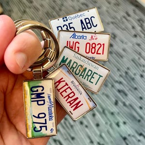 Può includere: Un portachiavi con cinque targhe in miniatura di diverse province canadesi. Le targhe sono dorate con lettere bianche e mostrano il nome della provincia e il numero di targa. Il portachiavi è attaccato a un anello argentato.