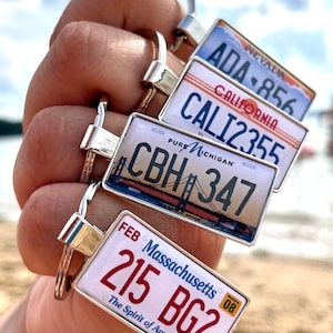May include: A keychain with five different license plate charms. The charms are from Nevada, California, Michigan, and Massachusetts. The charms are silver with colorful designs and text.