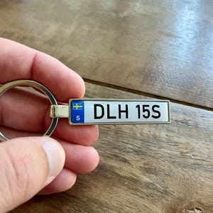 Puede incluir: Un llavero plateado con una etiqueta rectangular que se asemeja a una matrícula sueca. La etiqueta presenta el texto "DLH 15S" en negro, una bandera sueca azul y amarilla y la letra "S".