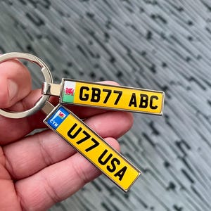 May include: A silver keychain with two yellow license plate charms. The first charm says "GB77 ABC" and has a small Welsh flag. The second charm says "U77 USA" and has a small Welsh flag.