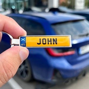 May include: A silver keyring with a yellow and blue Scottish flag charm and a yellow plate with the name "JOHN" on it.