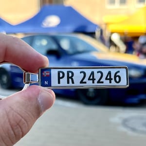 Peut inclure: Un porte-clés argenté avec un drapeau norvégien bleu et blanc et une plaque d'immatriculation argentée avec le texte "PR 24246".