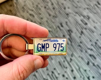 May include: A rectangular keychain featuring a miniature Manitoba license plate design. The plate displays "GMP 975" in blue against a white background, with "Friendly Manitoba" at the top. The keychain has a silver-colored ring.