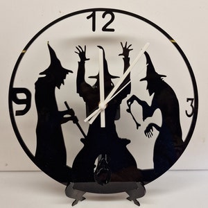 May include: Black and white clock featuring a silhouette of three witches stirring a cauldron. The clock face has Roman numerals with the numbers 3, 9, and 12 visible. The clock is on a black stand with three legs.