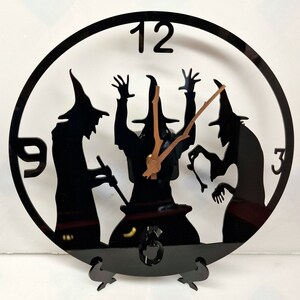 May include: Black clock with a silhouette of three witches stirring a cauldron. The clock face has Roman numerals and the numbers 9 and 3 are visible. The clock hands are gold.