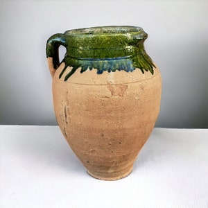 Może przedstawiać: Brązowy dzban ceramiczny z zieloną glazurą wokół górnej części. Dzban ma uchwyt i zaokrąglone ciało.