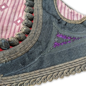 Puede incluir: Primer plano de una prenda tradicional con detalles intrincados. La pieza presenta una tela gris oscuro con patrones texturizados, acentuada por adornos marrones y elementos decorativos. Se ve una secci&oacute;n de tela roja y blanca estampada.