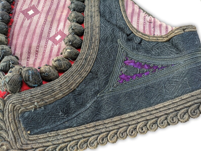 Puede incluir: Primer plano de una prenda tradicional con detalles intrincados. La pieza presenta una tela gris oscuro con patrones texturizados, acentuada por adornos marrones y elementos decorativos. Se ve una secci&oacute;n de tela roja y blanca estampada.