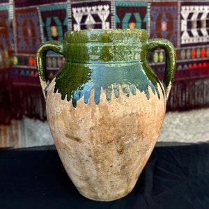 Pode incluir: Um vaso de cerâmica grande, de estilo antigo, com duas alças. A parte superior é esmaltada em verde escuro, com um efeito de gotejamento sobre o corpo bege. O vaso tem uma aparência rústica e envelhecida, sugerindo que é uma peça antiga. Altura aproximada 40 cm.
