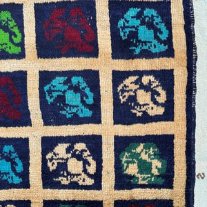 May include: A close-up of a rug with a repeating pattern of blue, green, and red stylized flowers on a beige background. The rug has a fringe border.