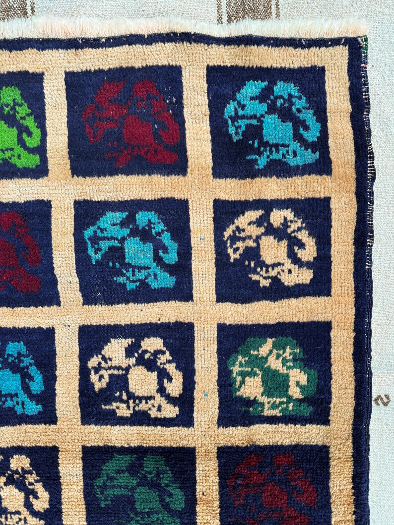 May include: A close-up of a rug with a repeating pattern of blue, green, and red stylized flowers on a beige background. The rug has a fringe border.