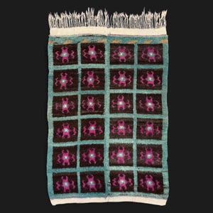 May include: Rectangular turquoise rug with a grid pattern of dark brown squares. Each square features a magenta and teal floral design. The rug has a white fringe along one edge, and is likely handmade.
