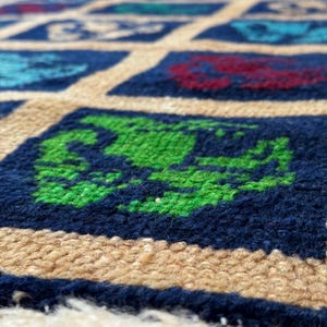 May include: Close-up of a blue and beige patterned rug with green, red, and blue squares. The rug has a fringe border.