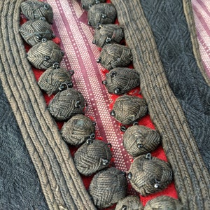 Puede incluir: Primer plano de un textil con detalles intrincados. El dise&ntilde;o presenta una tela gris oscuro con un patr&oacute;n de diamantes, bordeada por un ribete trenzado. Adornos redondos y tejidos est&aacute;n cosidos a lo largo de una secci&oacute;n a rayas rojas y blancas.