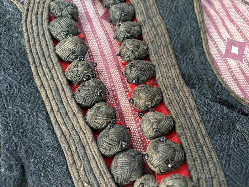 Puede incluir: Primer plano de un textil con detalles intrincados. El dise&ntilde;o presenta una tela gris oscuro con un patr&oacute;n de diamantes, bordeada por un ribete trenzado. Adornos redondos y tejidos est&aacute;n cosidos a lo largo de una secci&oacute;n a rayas rojas y blancas.