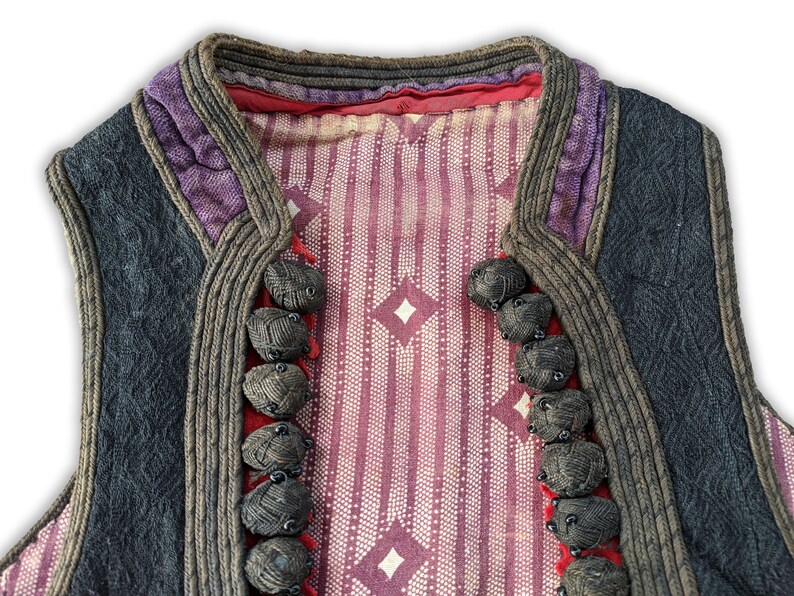 Puede incluir: Un chaleco vintage con un dise&ntilde;o estampado. El chaleco presenta un exterior gris oscuro con ribetes morados y marrones. El interior tiene un patr&oacute;n de rayas rojas y blancas con formas de diamante. Tiene varios botones oscuros en forma de bola gris.