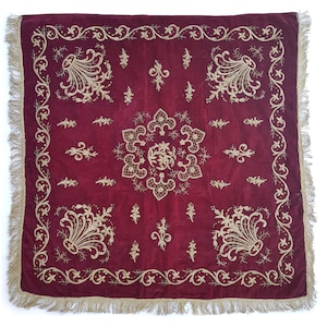 May include: A square, burgundy velvet cushion with a gold embroidered floral design. The cushion has a fringe border.