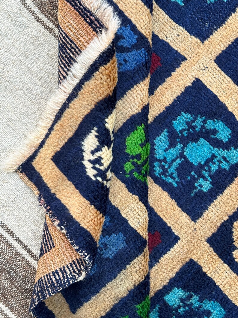 May include: A close-up of a colorful rug with a geometric pattern. The rug is made of wool and has a fringe border. The colors of the rug are blue, beige, green, and red.