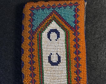 May include: A small, rectangular beaded pouch with a white background and a colorful geometric pattern. The pattern includes orange, red, green, blue, and white beads. The pouch has a traditional design with two crescent moon shapes in the center.