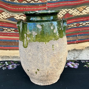 May include: A green glazed ceramic jar with two handles. The jar has a rough, textured surface and is partially covered in a green glaze. The jar is sitting on a black surface with a floral pattern.